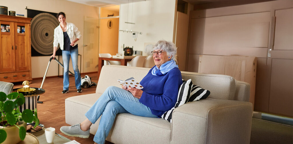 Oudere vrouw leest een tijdschrift op de bank terwijl een zorgmedewerker in de woonkamer stofzuigt.