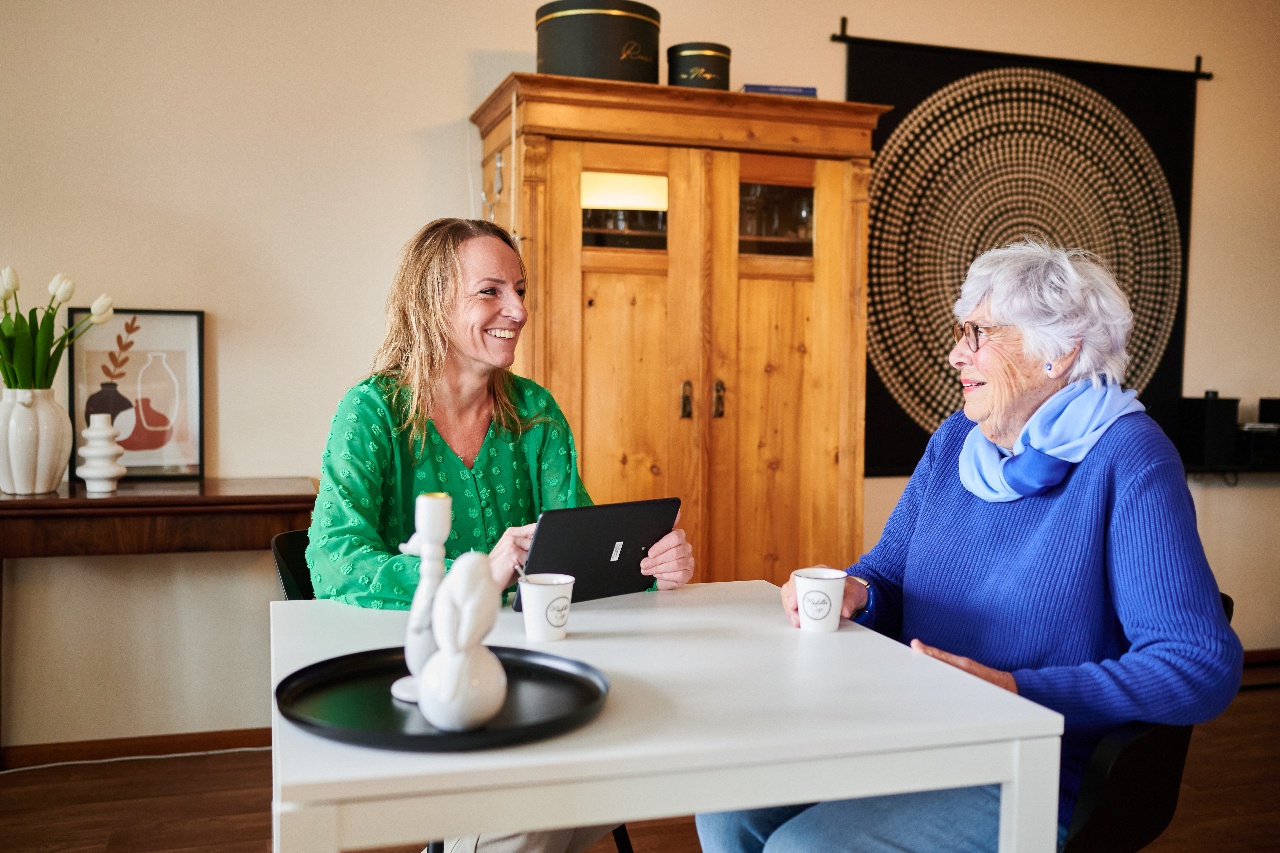 Oudere vrouw in blauwe trui zit lachend aan tafel tijdens een gesprek met een vrouw in groene blouse.