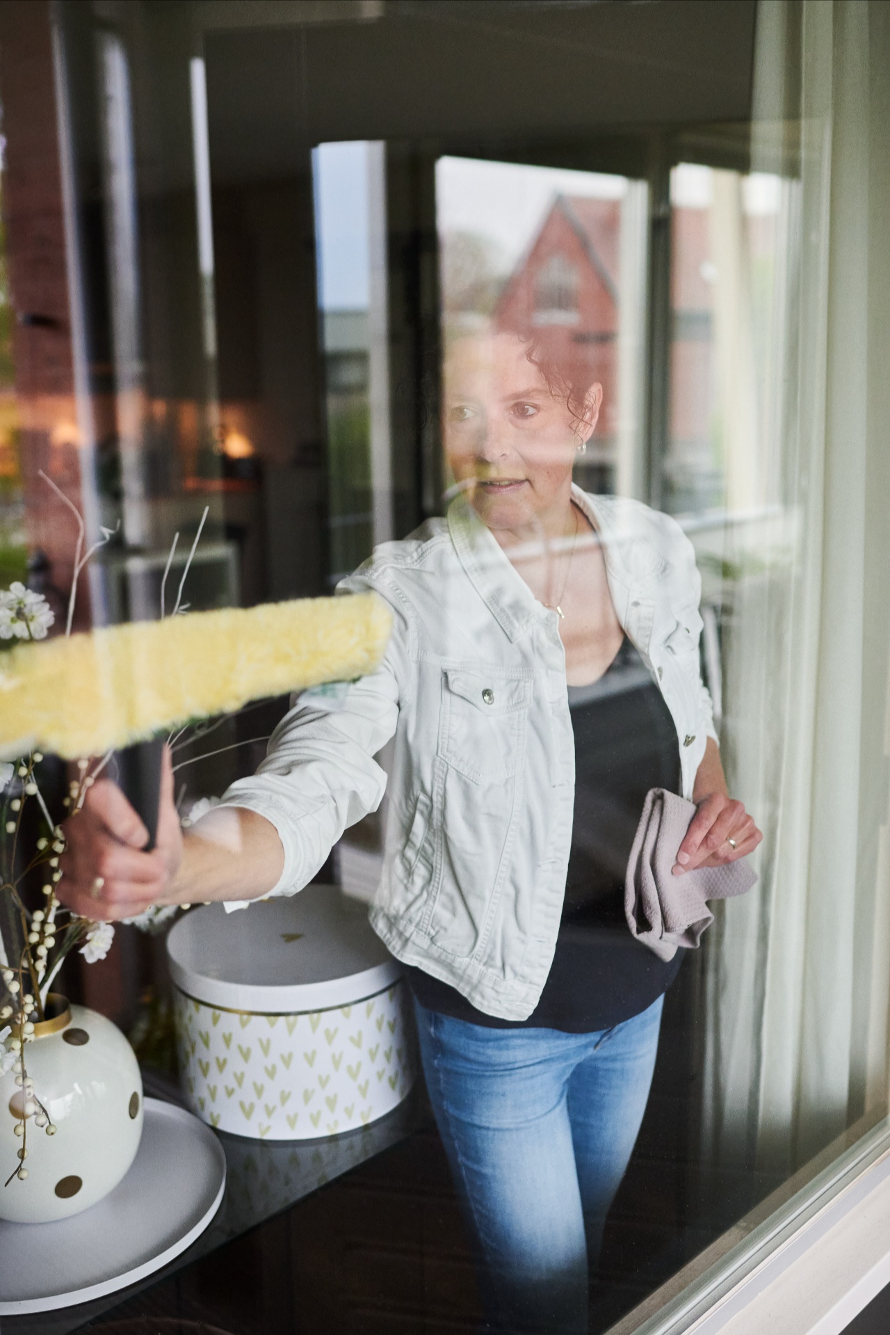Vrouw poetst een groot raam met een gele raamwisser terwijl ze glimlacht en geconcentreerd bezig is met schoonmaken.