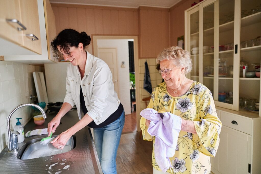 DNZT-medewerker wast af terwijl een oudere cliënt ernaast staat met een theedoek om te helpen afdrogen.