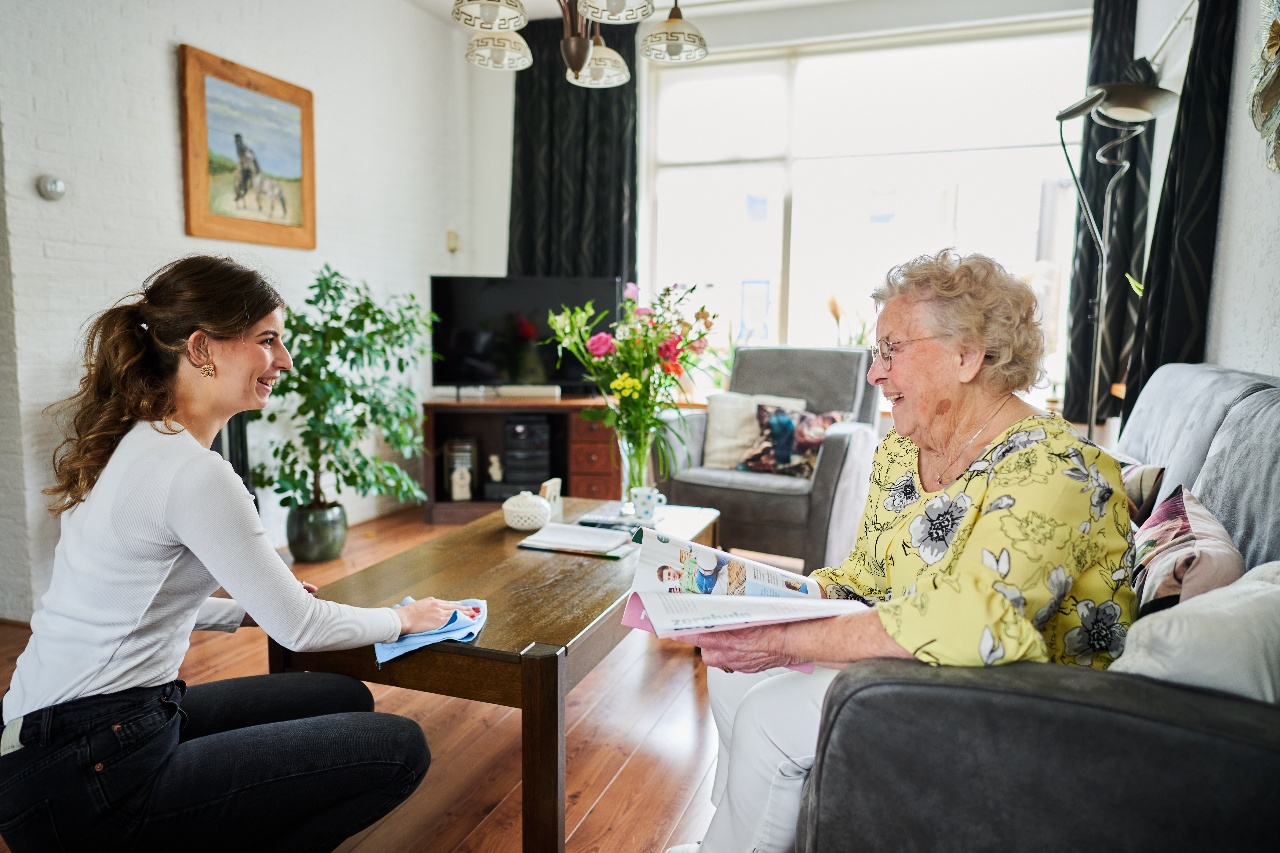 Oudere vrouw zit lachend op de bank in haar woonkamer met bloemen op tafel.