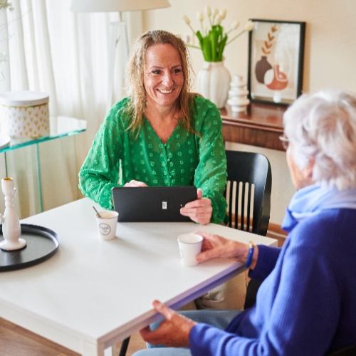 Vrouw in groene blouse in gesprek met oudere vrouw in blauwe trui aan tafel met tablet en koffiekopjes.