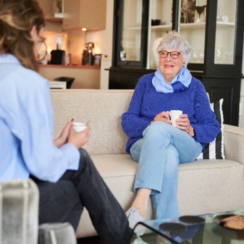 Vrouw in blauwe blouse drinkt koffie met een oudere vrouw in een woonkamer.